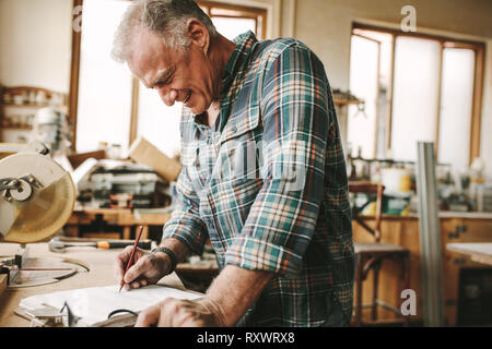 Happy senior carpenter preparing drawing for furniture parts. Man making designs on paper with manufacturing furniture. Stock Photo