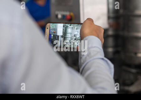 A man taking video with mobile phone the process of filling steel beer barrels with fresh beer in brewery plant factory Stock Photo