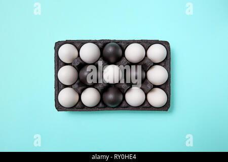 Pattern of white and painted black eggs in a black cardboard box on a blue background with copy space. Easter composition. Flat lay Stock Photo