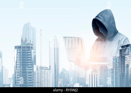 Double exposure of hacker in black hoodie holding laptop and modern city with skyscrapers background Stock Photo