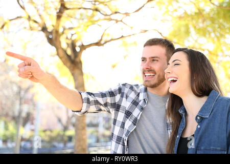 Happy couple pointing away laughing in a park with trees in the background Stock Photo