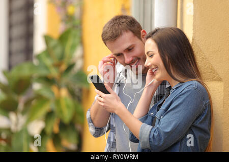Happy couple sharing earphones to watch media on smart phone in a colorful street Stock Photo