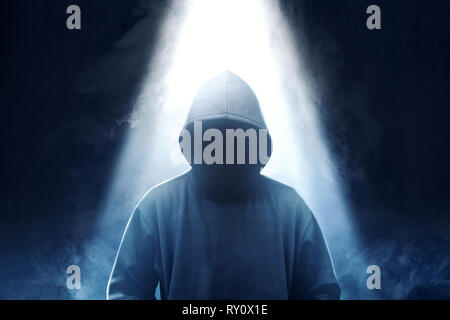 Hacker in black hoodie standing with smoke and light from the top over dark background Stock Photo