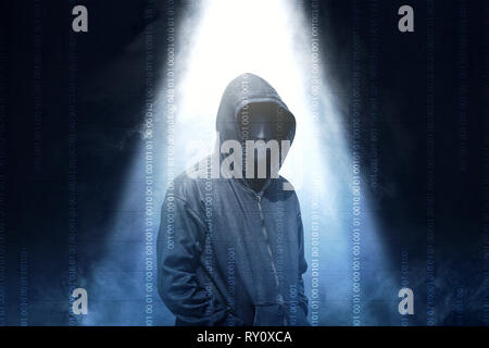 Hacker in black hoodie standing with smoke and light from the top over dark background Stock Photo