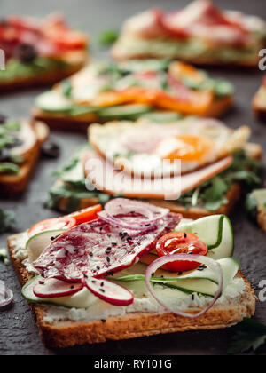 Close up view of different sandwiches with salami, vegetables and black sesame. Copy space for text. Assortment meat toasts on black background. Idea, creative concept for sausage maker. Vertical Stock Photo