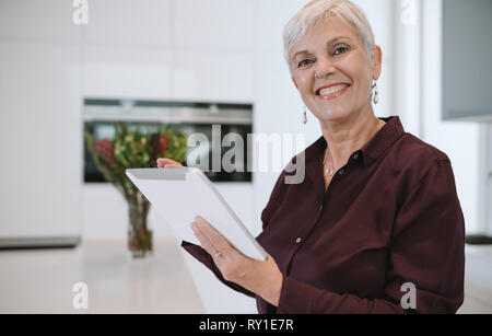 Portrait of a mature woman smiling, using her tablet at home , in the kitchen Stock Photo
