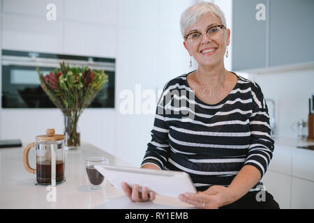 Portrait of a mature woman smiling wearing glasses, using her tablet at home , in the kitchen Stock Photo