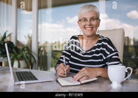 Beautiful senior woman using laptop at home. Mature smiling woman looking at camera while writing in notebook. Portrait of happy modern retired lady w Stock Photo