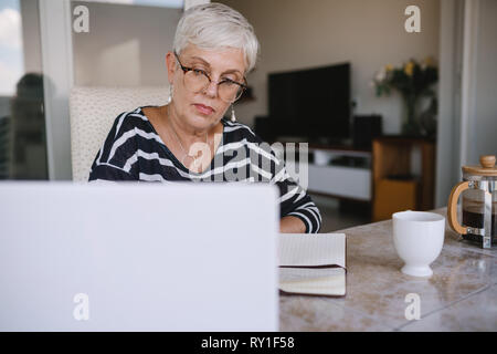 Mature beautiful woman making notes while working on her laptop at home. Elderly woman reading off her laptop while making notes Stock Photo