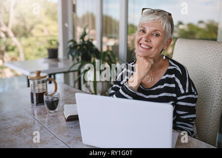 Beautiful senior woman using laptop at home. Mature smiling woman looking at camera while working with computer. Portrait of happy modern retired lady Stock Photo