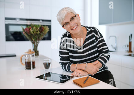 Portrait of a mature woman smiling wearing glasses and leaning on the kitchen counter. Senior woman working from home with tablet Stock Photo