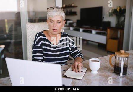 Mature beautiful woman working on her laptop at home. Elderly woman typing on her laptop while reading notes from her notebook outside on the balcony Stock Photo