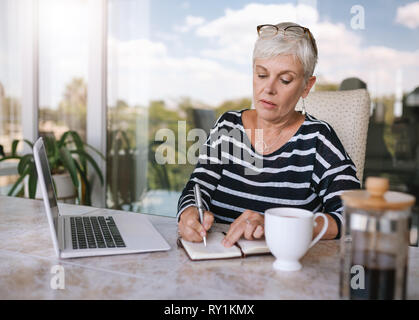 Mature beautiful woman making notes while working on her laptop at home. Elderly woman writing in her notebook outside on the balcony Stock Photo