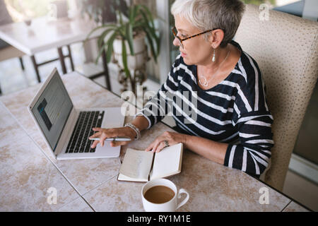 Mature beautiful woman working on her laptop at home. Elderly woman typing on her laptop while reading notes from her notebook outside on the balcony Stock Photo