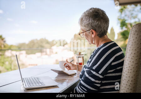 Happy modern retired lady with laptop and notebook sitting outside on balcony. Elderly woman working from home outside on her balcony Stock Photo