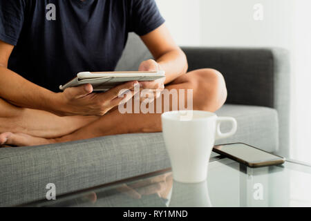 closeup of a young caucasian man in shorts sitting, cross-legged, in a sofa while is using a tablet, and a cup of coffee and a smartphone on a glass c Stock Photo