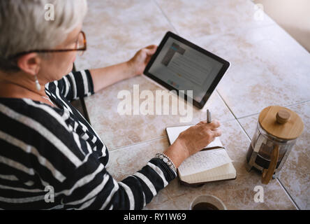 Top view of mature woman making notes in her notebook or journal while reading off the screen of a digital tablet Stock Photo