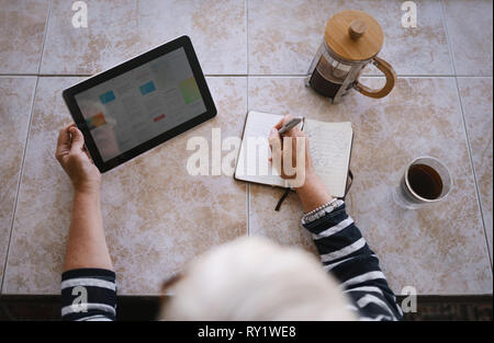 Top view of mature woman making note in her notebook or journal while reading off an article on a digital tablet Stock Photo