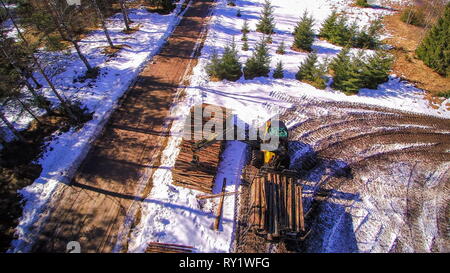 The aerial view of the wide snowy forest ground with the piles of logs on the ground being grabbed by the forest harvester Stock Photo