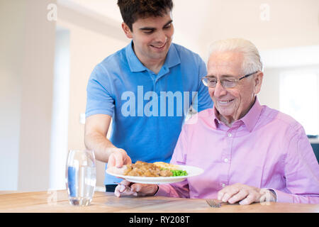 Male Care Assistant Serving Meal To Senior Male Seated At Table Stock Photo