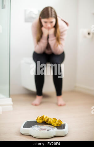 Unhappy Teenage Girl Sitting In Bathroom Looking At Scales And Tape Measure Stock Photo
