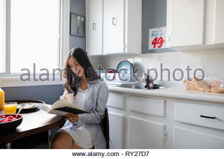Young Latinx woman drinking coffee and reading book in morning kitchen