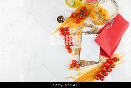 Notebook on wooden cutting board with different types of dry pasta and tomatoes on white wooden background, top view. Copy space. Flat lay. Stock Photo