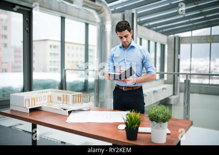 Young businessman or architect with model of a house standing at the desk in office, making notes when working. Stock Photo