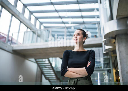 A waist-up portrait of young businesswoman standing in corridor outside office, arms crossed. Stock Photo