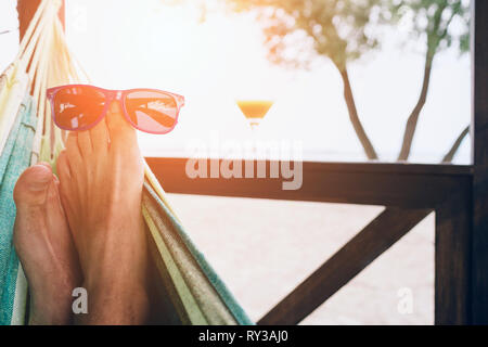 Young man lying in a hammock at a sunny beach by ocean. Sunglasses on the feet. Stock Photo