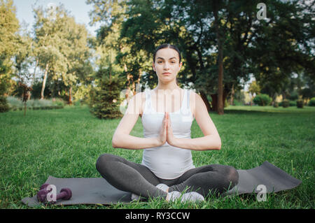 Young pregnant woman meditating outside. She sit on yoga mate in lotus pose. Model keep hands in praying position. Stock Photo