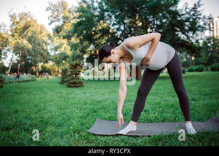 Active young pregnant woman exercising on yoga mate in park. She reach ground with one hand and hold another on belly. Stock Photo