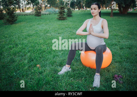 Young pregnant woman sit on orange fitness ball in green park and meditate. She hold hands together in praying pose. Stock Photo