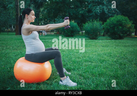 Picture of nice young pregnant woman sit on orange fitness ball in green park. She exercise with dumbells. Model look concentrated. Stock Photo