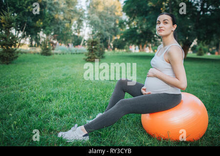 Pretty young pregnant woman sit on orange fitness ball in park. She hold hands around belly and look at it. Stock Photo