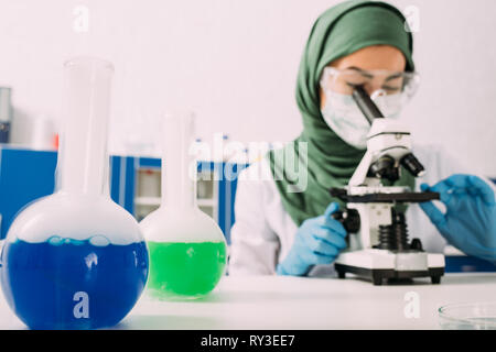 female muslim scientist sitting at table with flasks and using microscope during experiment in chemical laboratory Stock Photo