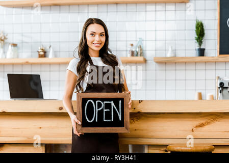 attractive cashier in brown apron standing near wooden bar counter and holding chalkboard with open lettering in coffee house Stock Photo