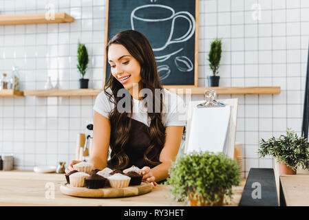 attractive waitress standing behind wooden bar counter and holding tray with cupcakes in coffee house Stock Photo