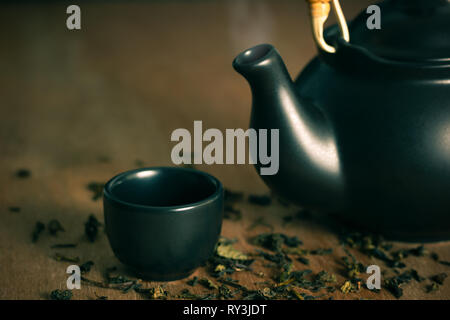 Glass of tea with hot smoke is placed beside a ceramic teapot and dried tea leaves scattered on the wood background. Stock Photo