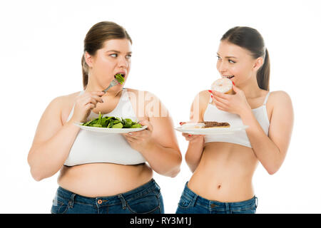 slim woman eating doughnuts and overweight woman eating green spinach leaves while looking at each other isolated on white Stock Photo