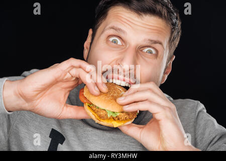 emotional man eating tasty chicken burger while looking at camera isolated on black Stock Photo