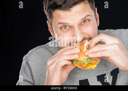 man eating tasty chicken burger while looking at camera isolated on black Stock Photo