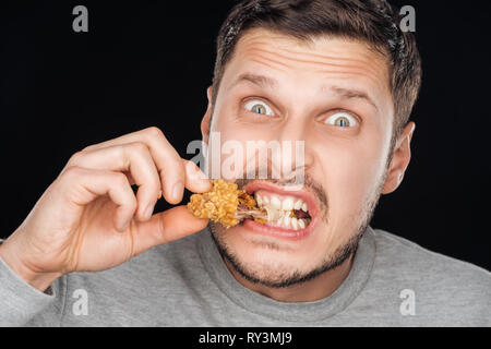 emotional man eating crispy chicken nugget while looking at camera isolated on black Stock Photo