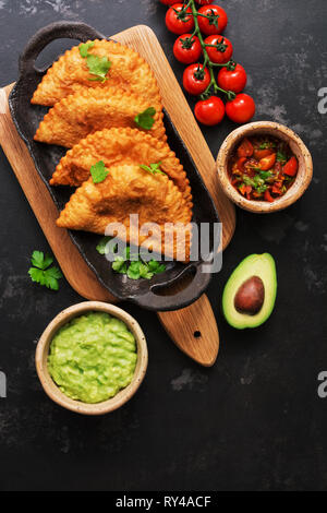Latin American, Mexican, Chilean food. Traditional fried empanadas served with tomato and avocado sauce on a dark background. Top view, flat lay.