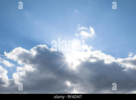 The sun's rays come out from behind the clouds in the bright blue sky. Stock Photo