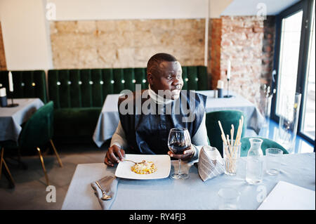 African man in black traditional clothes sitting at restaurant and eat pasta and drink wine. Stock Photo