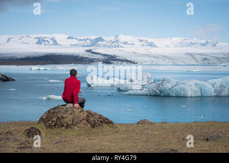 Man traveler in red jacket sits on the shore of a lake. Summer landscape with glacial lagoon, glacier and icebergs in the southeast of Iceland, Europe Stock Photo