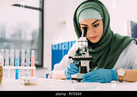 beautiful female muslim scientist looking through microscope during experiment in chemical laboratory Stock Photo