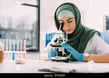 beautiful female muslim scientist looking through microscope during experiment in chemical laboratory Stock Photo