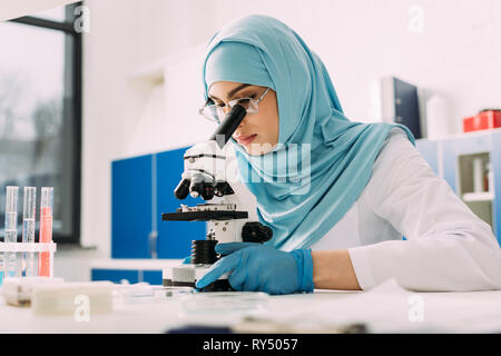 concentrated female muslim scientist looking through microscope during experiment in chemical laboratory Stock Photo